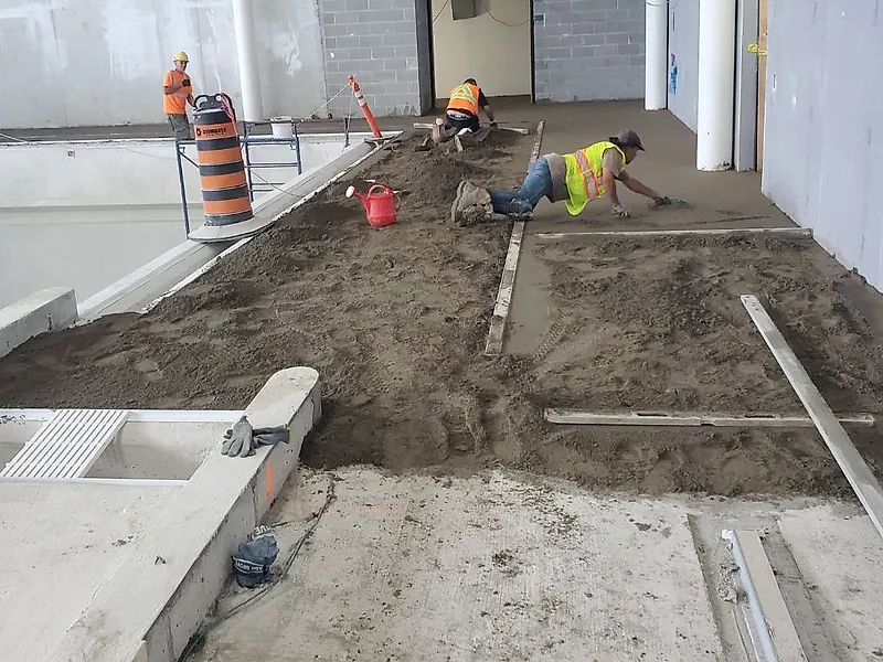 Three workers on an indoor pool deck, one lying flat to float the mortar, with screed rails and loose mortar around them